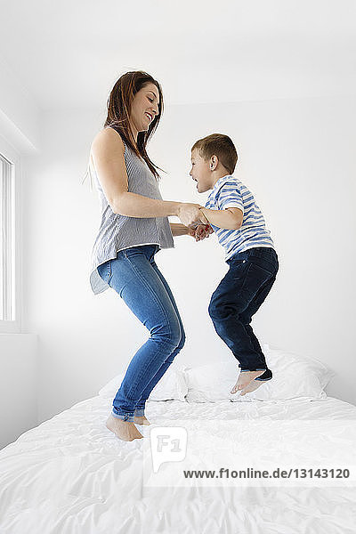 Verspielte Mutter und Sohn halten sich in voller Länge an den Händen  während sie zu Hause auf dem Bett hüpfen