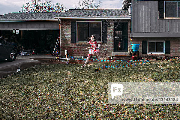 Verspieltes Mädchen rennt im Garten im Sprinkler gegen das Haus