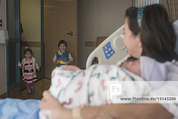 Mutter hält neugeborenen Sohn und sieht Töchter  die in die Krankenhausstation kommen  während sie sich auf dem Bett ausruht