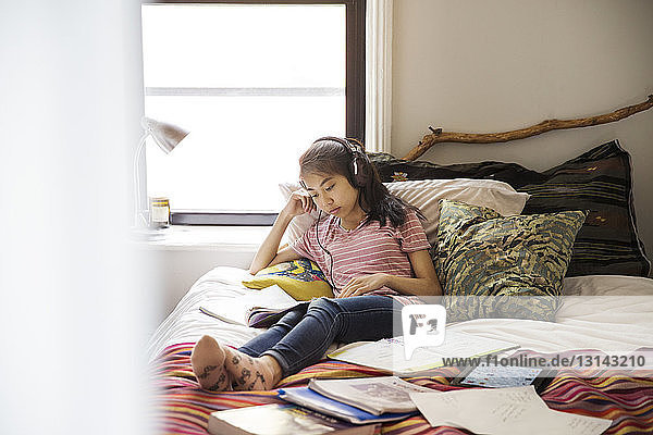 Teenage girl with headphones studying while sitting on bed