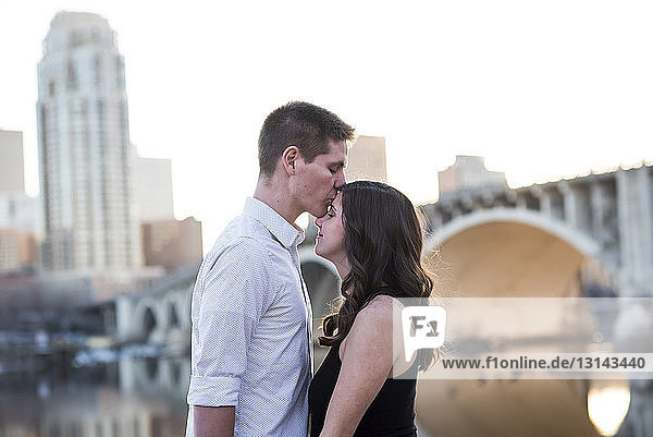 Husband kissing on pregnant wife's forehead while standing by river against clear sky in city