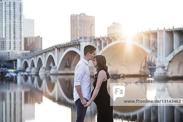 Husband kissing on pregnant wife's forehead while standing by river against clear sky