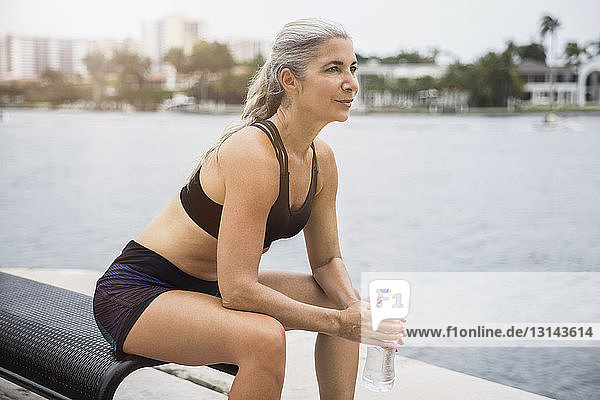 Nachdenkliche  sportliche Frau  die eine Wasserflasche hält  während sie auf einer Bank am Fluss sitzt