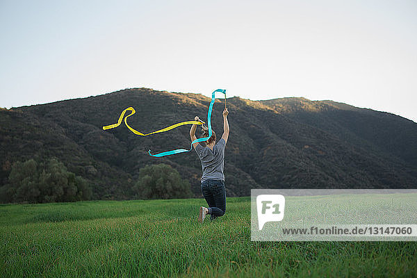 Rear view of young woman running in field holding up dance ribbons