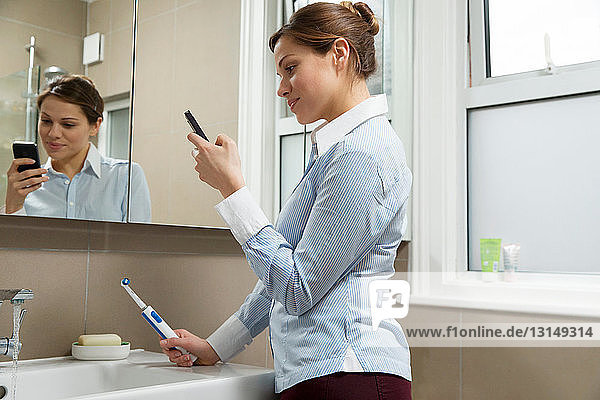 Young woman using cell phone holding toothbrush