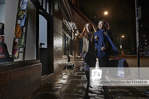 Vorderansicht eines Paares  das Arm in Arm eine Straße entlangläuft und eine Einkaufstasche bei Nacht trägt  Schaufensterbummel