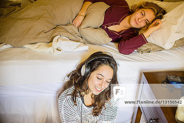 Two young female friends lying in bed and listening to headphones