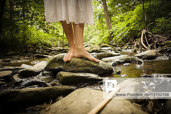 Bare feet of mid adult woman wearing white dress balancing on rock in riverbed