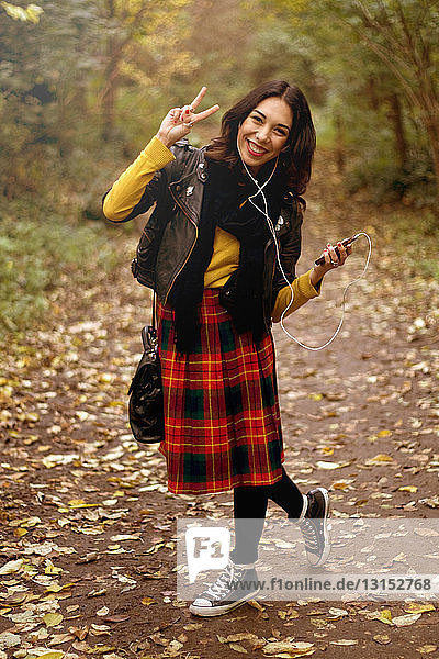 Young woman in park  listening to music using earphones  making peace sign