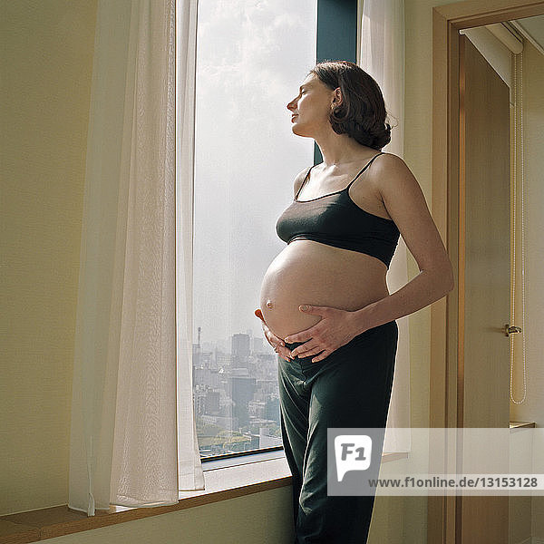 Porträt einer mittelgroßen schwangeren Frau  die am Wohnungsfenster steht und den Bauch hält