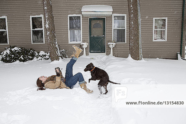 Mittlere erwachsene Frau liegt im Schnee vor dem Haus und spielt mit einem Hund