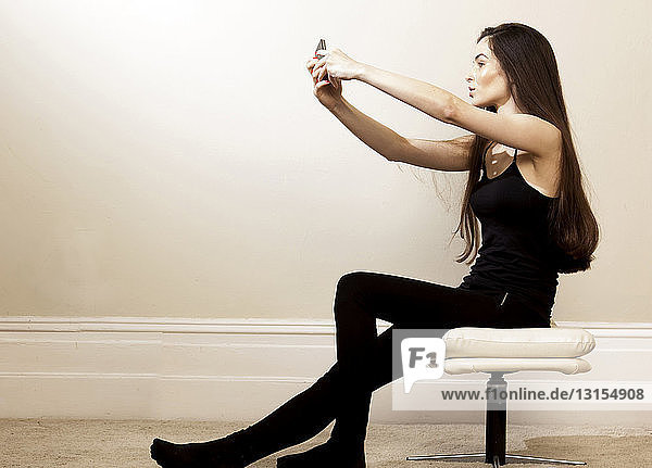 Seitenansicht einer jungen Frau  die auf einem Hocker sitzt und ein Smartphone benutzt  um ein Selfie zu machen