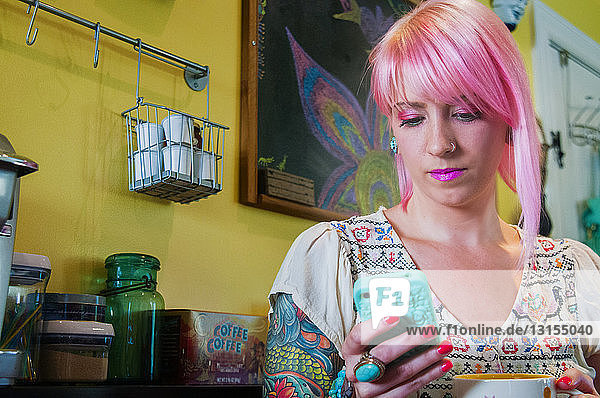 Junge Frau mit rosa Haaren liest in der Küche eine Textnachricht auf dem Smartphone