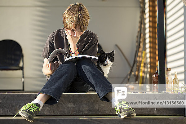 Junge sitzt auf einer Stufe und liest ein Buch  die Katze schaut von hinten zu