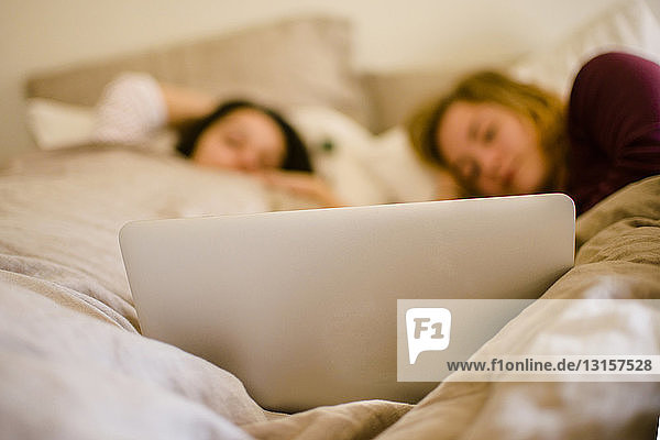 Two young female friends lying in bed looking at laptop