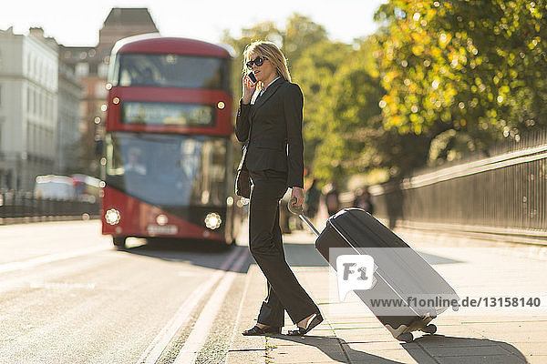 Junge Geschäftsfrau mit Rollkoffer  die mit ihrem Smartphone spricht  London  Vereinigtes Königreich