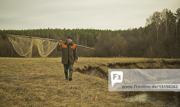 Mature man walking along riverbank carrying traditional fishing net