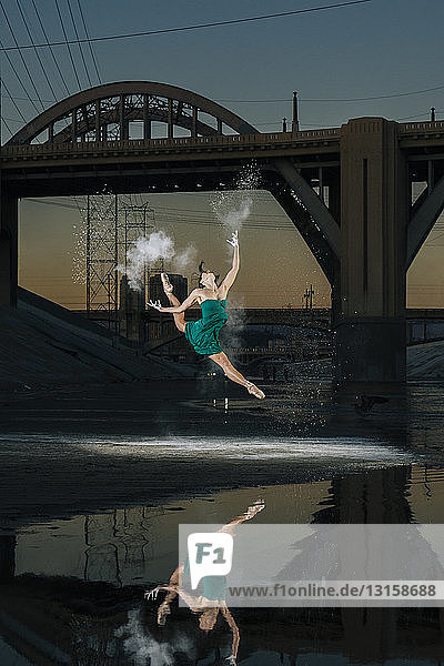 Weibliche Balletttänzerin springt  während sie eine Pulverexplosion über einem Fluss bei Sonnenuntergang auslöst  Los Angeles  USA