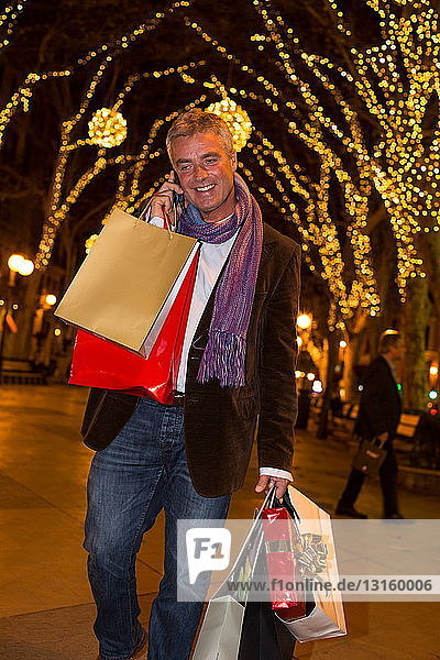 Älterer Mann mit Weihnachtseinkäufen  der auf einer von Bäumen gesäumten Allee mit seinem Smartphone spricht  Mallorca  Spanien