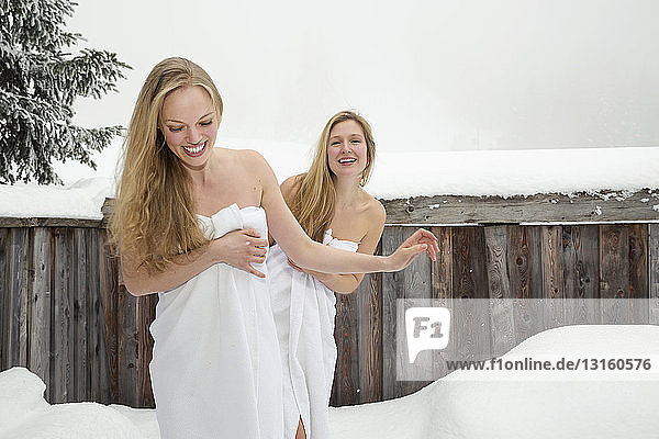 Two young women wrapped in white towels outside log cabin sauna