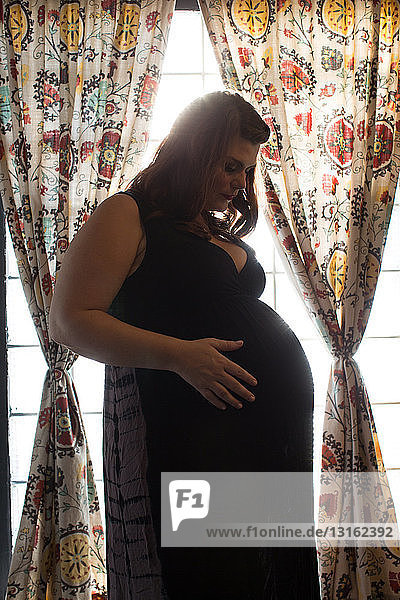 Seitenansicht einer schwangeren Frau mittleren Alters  die vor dem Fenster steht und nach unten schaut und den Bauch berührt