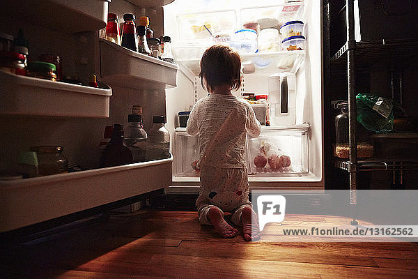 Rückansicht eines männlichen Kleinkindes  das nachts vor einem offenen Kühlschrank kniet