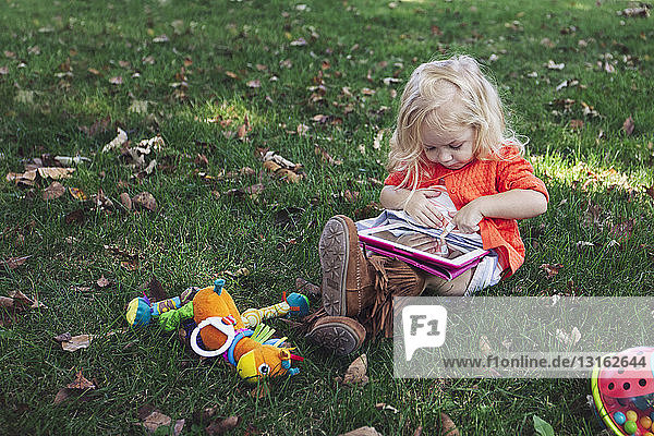 Mädchen sitzt mit Spielzeug im Gras und schaut mit einem digitalen Tablett nach unten
