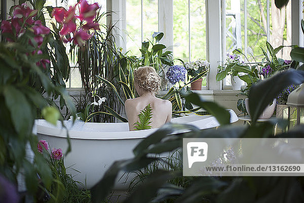 Nackte junge Frau  in Badewanne sitzend  im Badezimmer voller Pflanzen  Rückansicht
