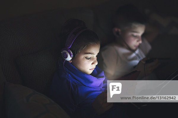 Boy and girl side by side on sofa wearing earphones using digital tablets