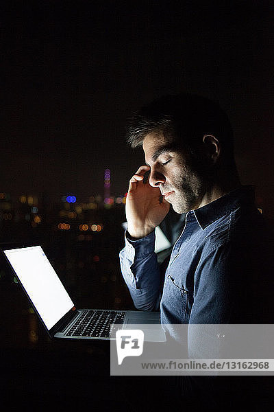 Müde junger Geschäftsmann mit Laptop vor dem Fenster eines nächtlichen Bürohochhauses  Shanghai  China