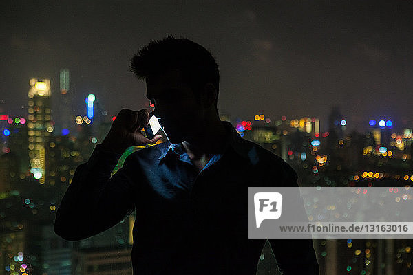Silhouettierter junger Geschäftsmann plaudert nachts vor dem Fenster eines Hochhaus-Büros mit seinem Smartphone  Shanghai  China