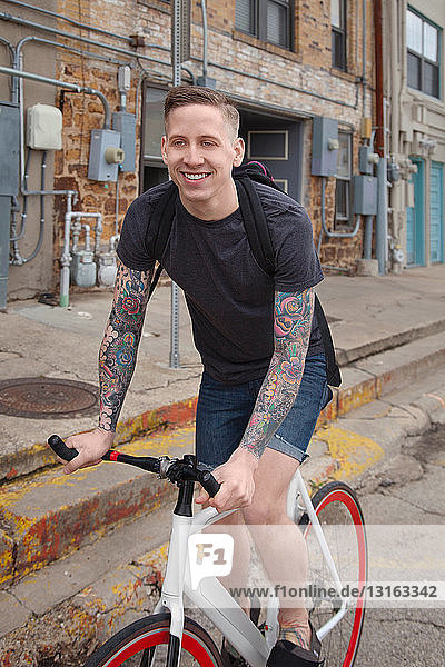 Junger tätowierter Mann fährt mit dem Fahrrad auf der Straße und lächelt