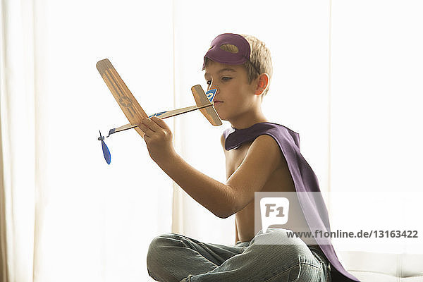 Junge mit Umhang und Maske spielt zu Hause mit Spielzeugflugzeug