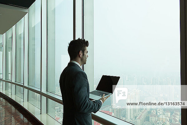 Junger Geschäftsmann mit Laptop  der aus dem Fenster eines Bürohochhauses schaut  Shanghai  China