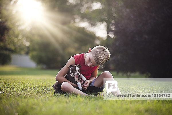 Frontansicht eines Jungen  der mit einem Boston-Terrier-Welpen im Gras sitzt und nach unten schaut