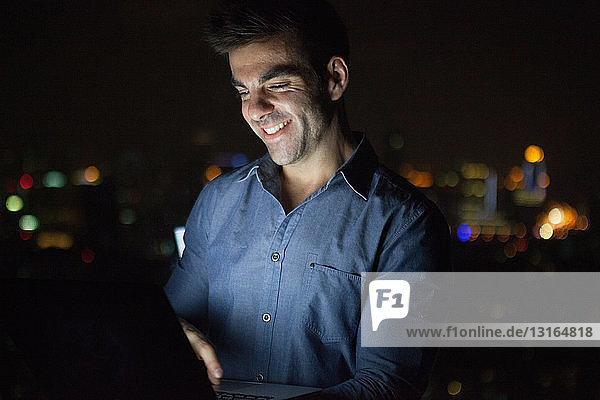 Junger Geschäftsmann tippt nachts vor dem Fenster eines Bürohochhauses auf einer Laptop-Tastatur  Shanghai  China