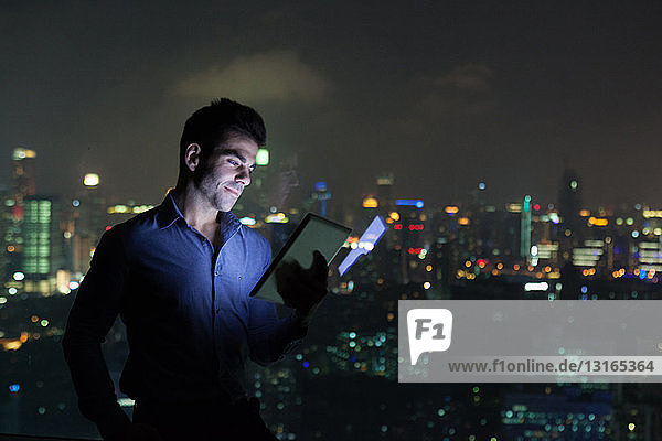 Junger Geschäftsmann betrachtet digitales Tablet vor dem Fenster eines Bürohochhauses bei Nacht  Shanghai  China