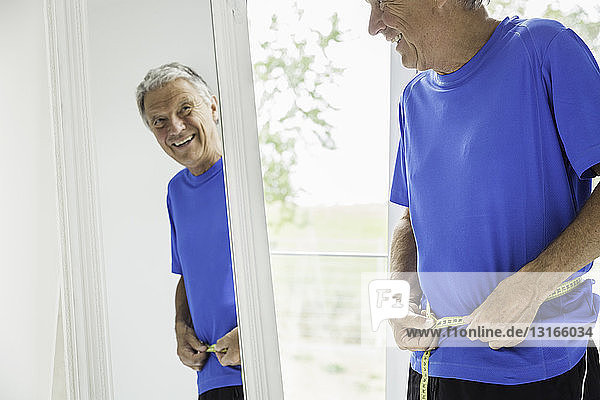 Glücklicher älterer Mann misst seine Taille  während er in den Spiegel schaut
