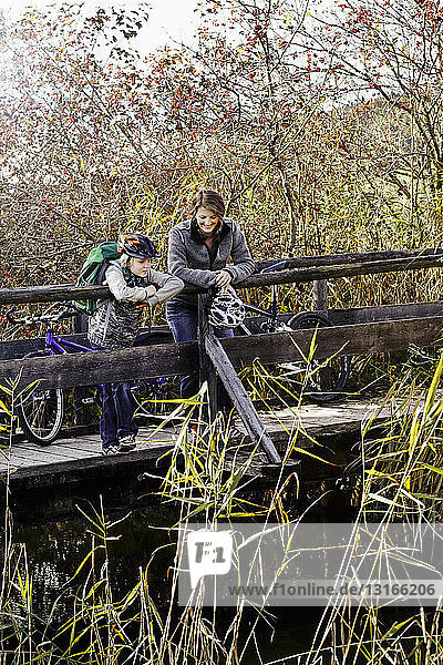 Mother and son with bicycles standing on wood footbridge looking down at river