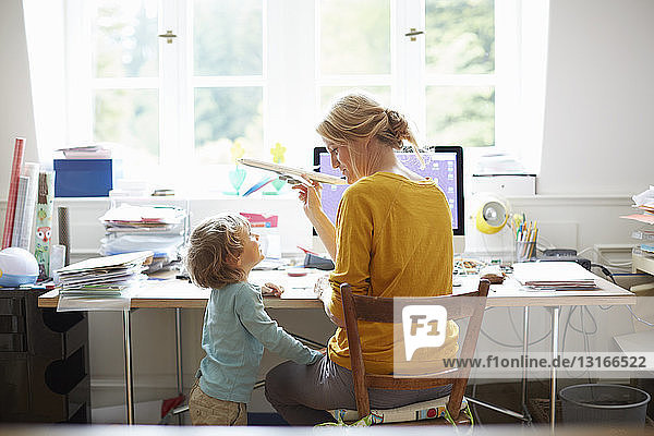Rear view of mature woman and boy sitting at computer playing with toy aeroplane