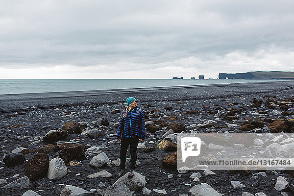 Mittelgroße erwachsene Frau an felsiger Küste stehend  wegschauend  Island