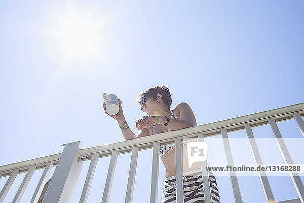 Niedrigwinkelansicht einer mittelgroßen erwachsenen Frau  die mit einem Kaffeebecher am Balkon lehnt
