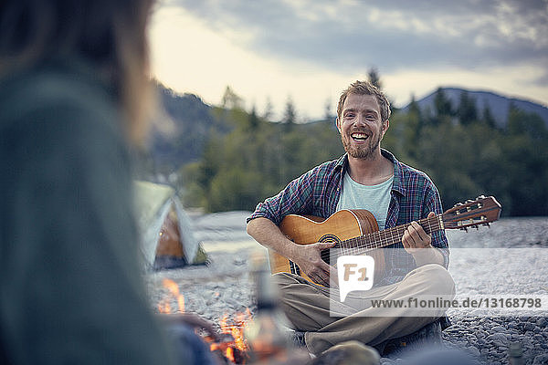 Frontansicht eines jungen Mannes  der am Lagerfeuer sitzt und Gitarre spielt