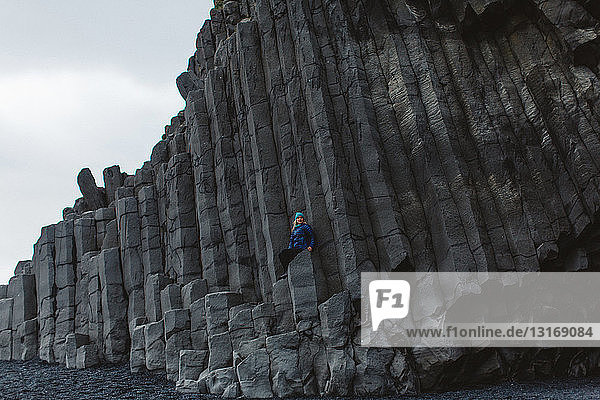 Mittelgroße erwachsene Frau sitzt auf einer säulenförmigen Basaltfelswand und schaut in die Kamera  Island