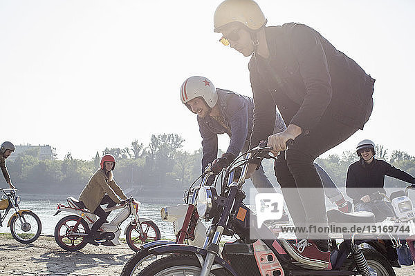 Gruppe von Freunden fährt mit Moped am See  zwei Männer machen Stunts