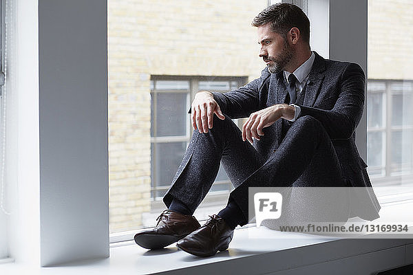 Portrait of businessman sitting on windowsill looking out of window