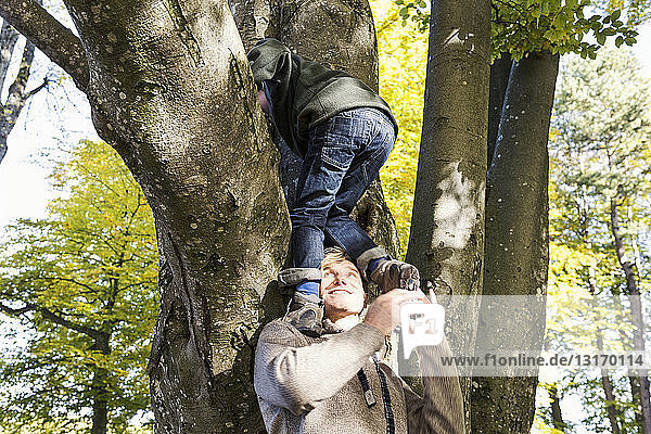 Vater hilft Sohn beim Baumklettern und schaut lächelnd auf