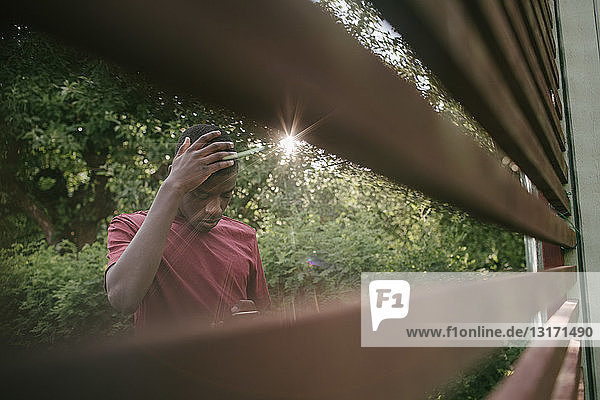 Junge berührt den Kopf während er ein Mobiltelefon benutzt  gesehen durch einen Zaun im Hinterhof