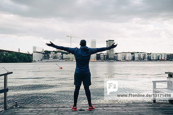 Rückansicht eines männlichen Athleten  der mit ausgestreckten Armen auf dem Pier über dem Meer in der Stadt steht