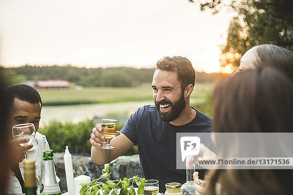 Cheerful male and female friends enjoying drink dinner in backyard during sunset